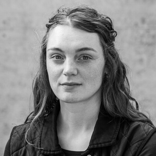 Великий план, чорно-білий портрет. Дівчина з довгим темним хвилястим волоссям, темними бровами й світлими очима дивиться в кадр і майже непомітно всміхається. На ній темні футболка й куртка.