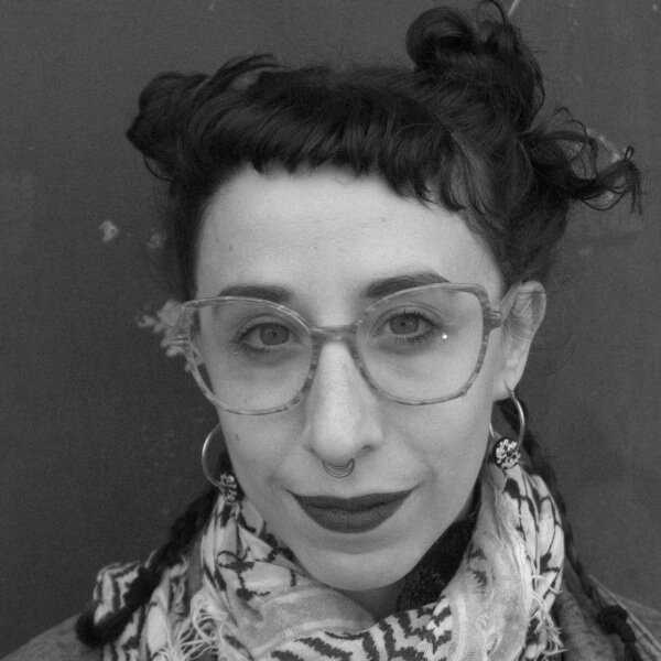 Великий план, чорно-білий портрет. Жінка уважно дивиться в камеру. Її темне волосся зібране в дві високі гульки, на плечах лежать дві тонесенькі косички, гривка підстрижена дуже коротко. На жінці великі окуляри в легкій оправі, круглі сережки, куфія. Нафарбовані насиченою помадою губи злегка всміхаються.