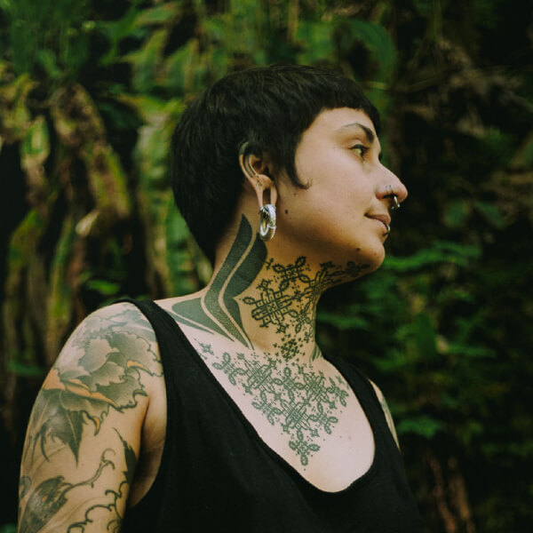 Погрудний план, портрет. На тлі рясної зелені дерев людина з коротким чорним волоссям, виразними бровами, стоїть боком до камери. Вона вдягнена у чорну майку. Її шию, груди та плечі прикрашають татуйовання, а вуха і ніс – пірсинг.