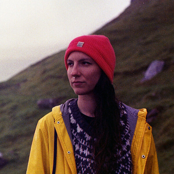 Погрудний план, портрет. На тлі туманного моря й зеленкуватого схилу людина дивиться вдалечінь. У неї довге темне волосся, в носі пірсинг. На ній червона шапка, в’язаний светр, жовтий дощовик.