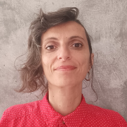 Великий план, портрет. Жінка з асиметрично підстриженим каштановим волоссям, темними бровами й очима дивиться у камеру з легкою усмішкою. На ній червона крапчаста сорочка, у лівому вусі – кругла сережка. Позаду – сіре тло.